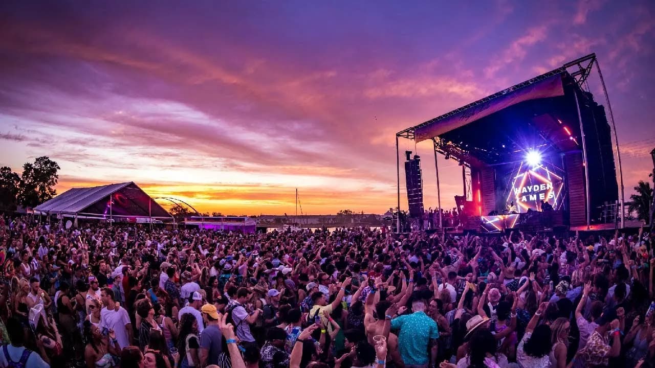 Festival crowd dancing at sunset in front of an outdoor stage during a Hayden James set