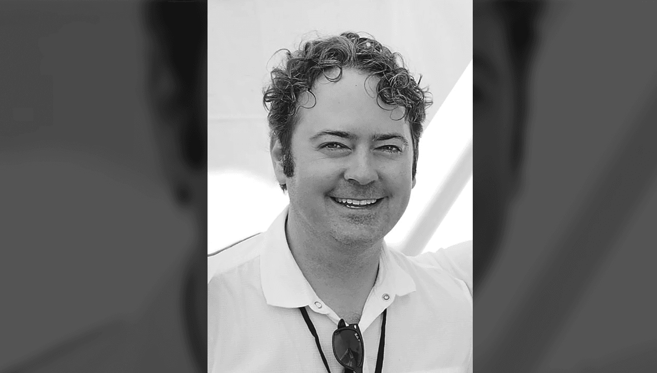 Black-and-white portrait of JK McKnight smiling in a white polo shirt with sunglasses hanging from the collar.