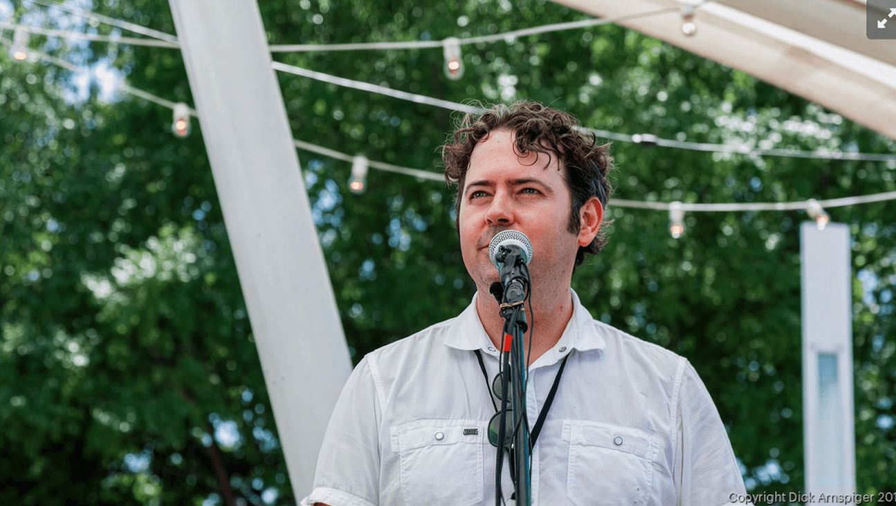 JK McKnight speaking into a microphone at an outdoor festival, wearing a white shirt beneath a tent canopy strung with lights and framed by green trees.