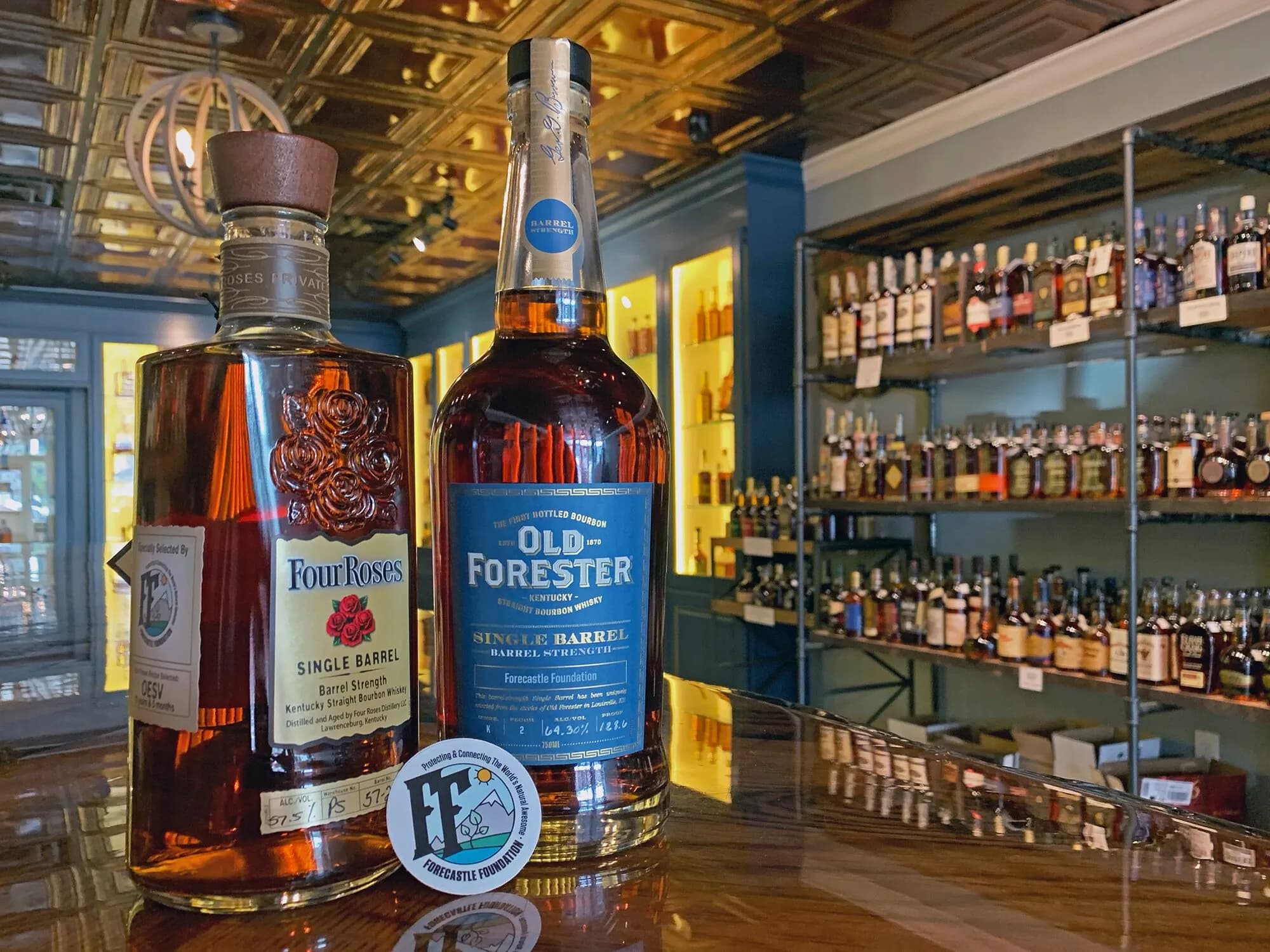 Four Roses and Old Forester single barrel bourbon bottles labeled for the Forecastle Foundation, displayed on a bar beside a Forecastle Foundation sticker