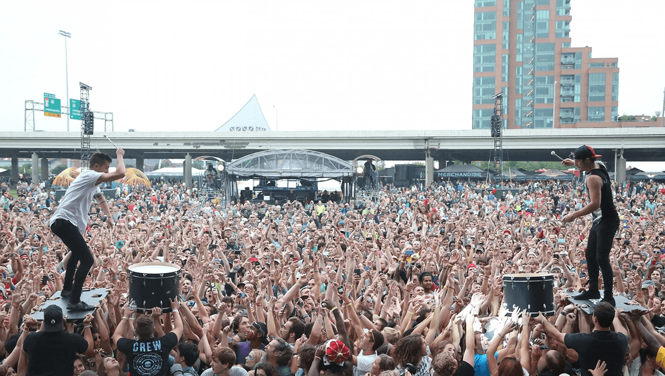 Two drummers performing on platforms above a massive Forecastle Festival crowd with thousands of hands raised, the Louisville skyline behind the stage.