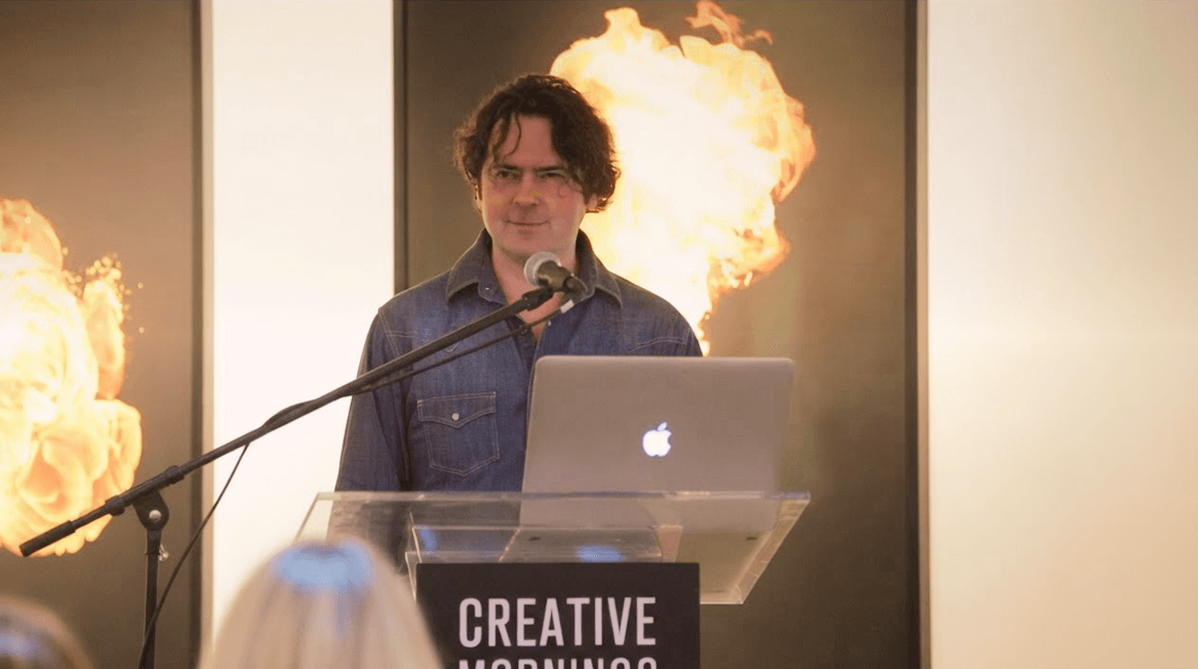 JK McKnight speaking at a CreativeMornings event, standing at a clear podium with a laptop as fireballs erupt on the stage behind him.