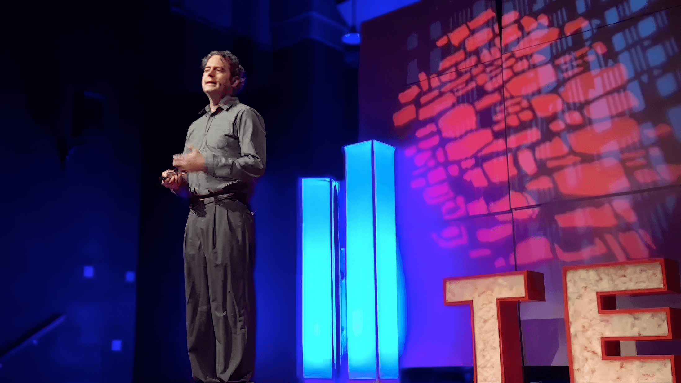 JK McKnight speaking on stage at a TED event, with glowing blue columns and red TED lettering behind him.
