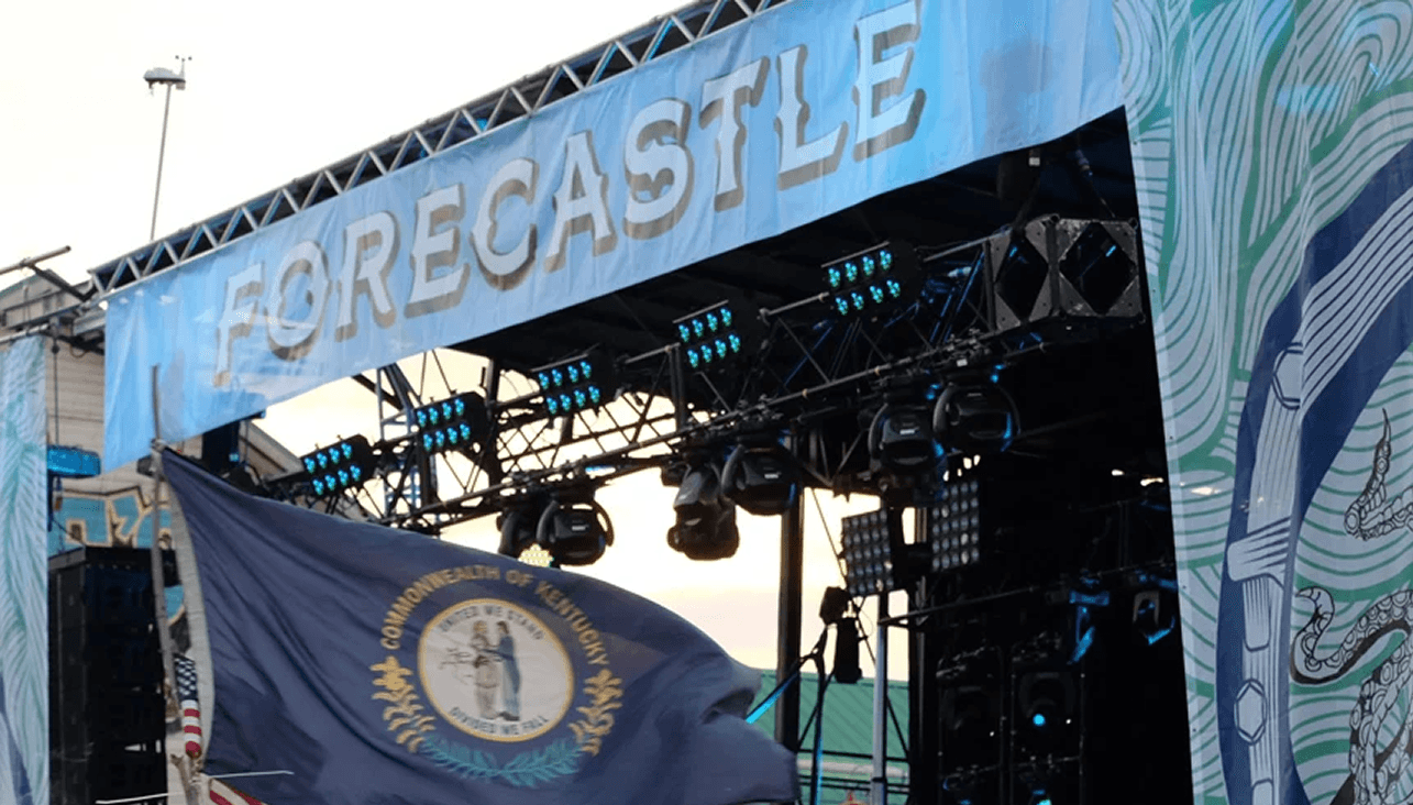 The Forecastle Festival main stage at dusk with a large FORECASTLE banner across the top truss and a Commonwealth of Kentucky flag waving in the foreground.