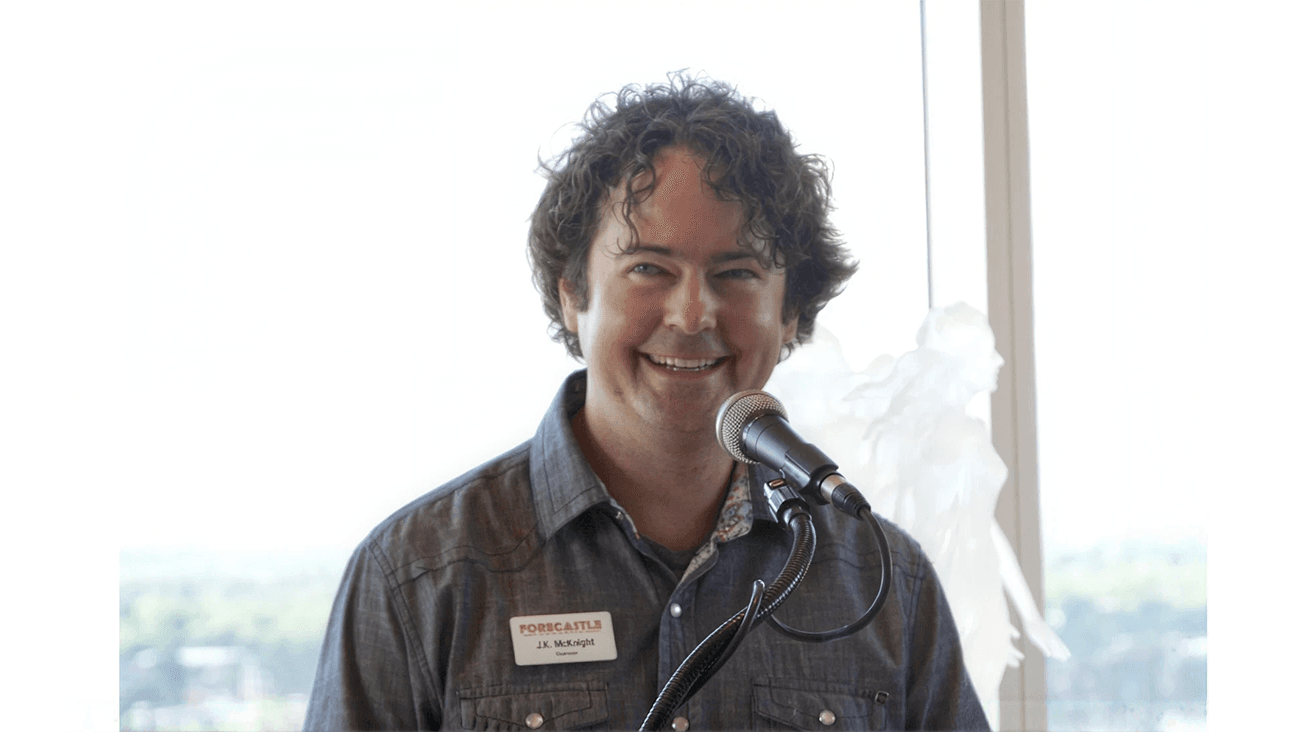 JK McKnight smiling behind a microphone in a gray denim shirt with a Forecastle chairman name badge, in front of a bright window overlooking a hazy skyline.