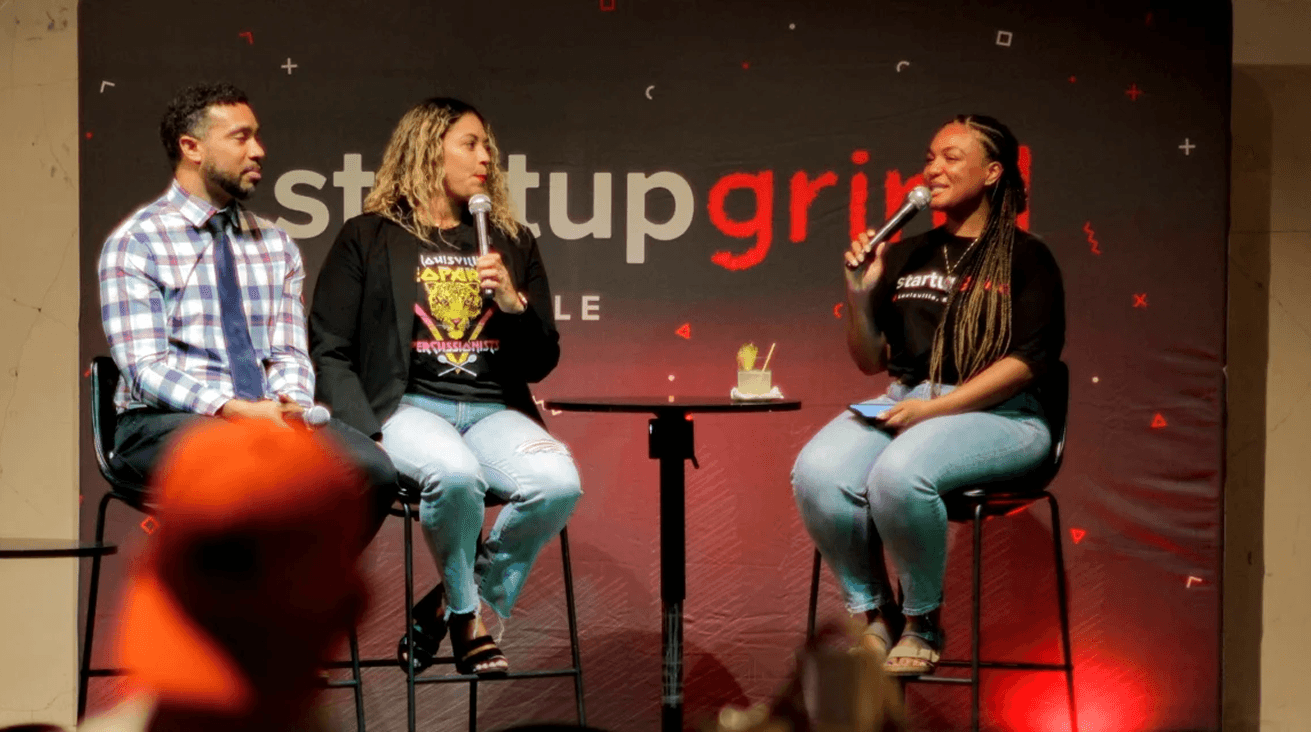 Three panelists seated on stools on stage during a Startup Grind Louisville fireside chat, two holding microphones in front of the red Startup Grind backdrop.