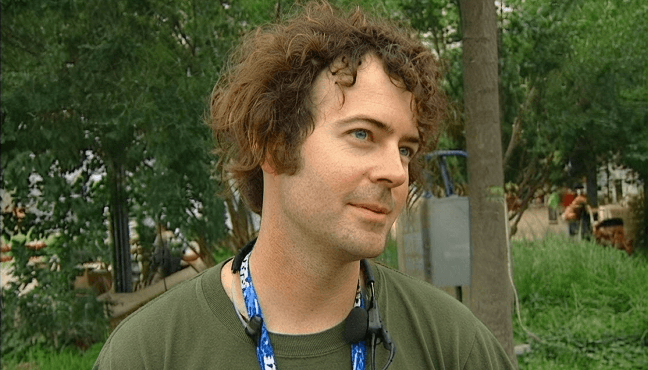 JK McKnight being interviewed outdoors in an olive-green t-shirt with a festival lanyard and clip-on mic, green trees and grass behind him.