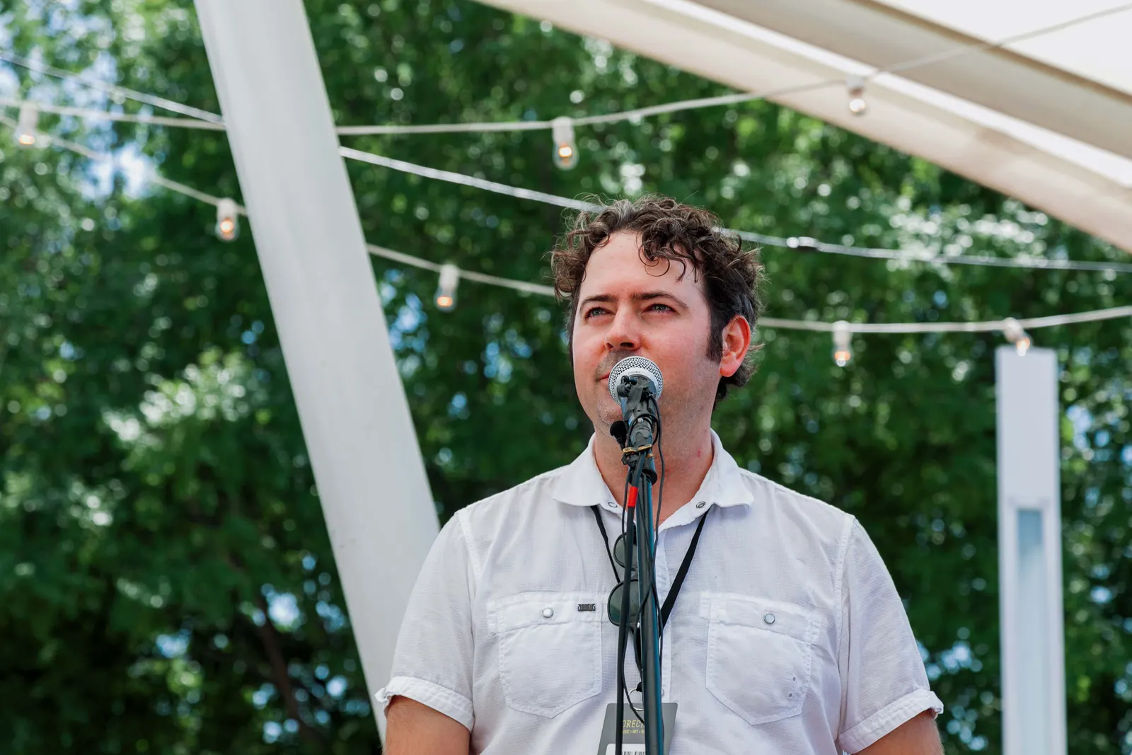 JK McKnight speaking into a microphone outdoors in a white short-sleeve button-up shirt with a festival lanyard, green trees and string lights behind him.