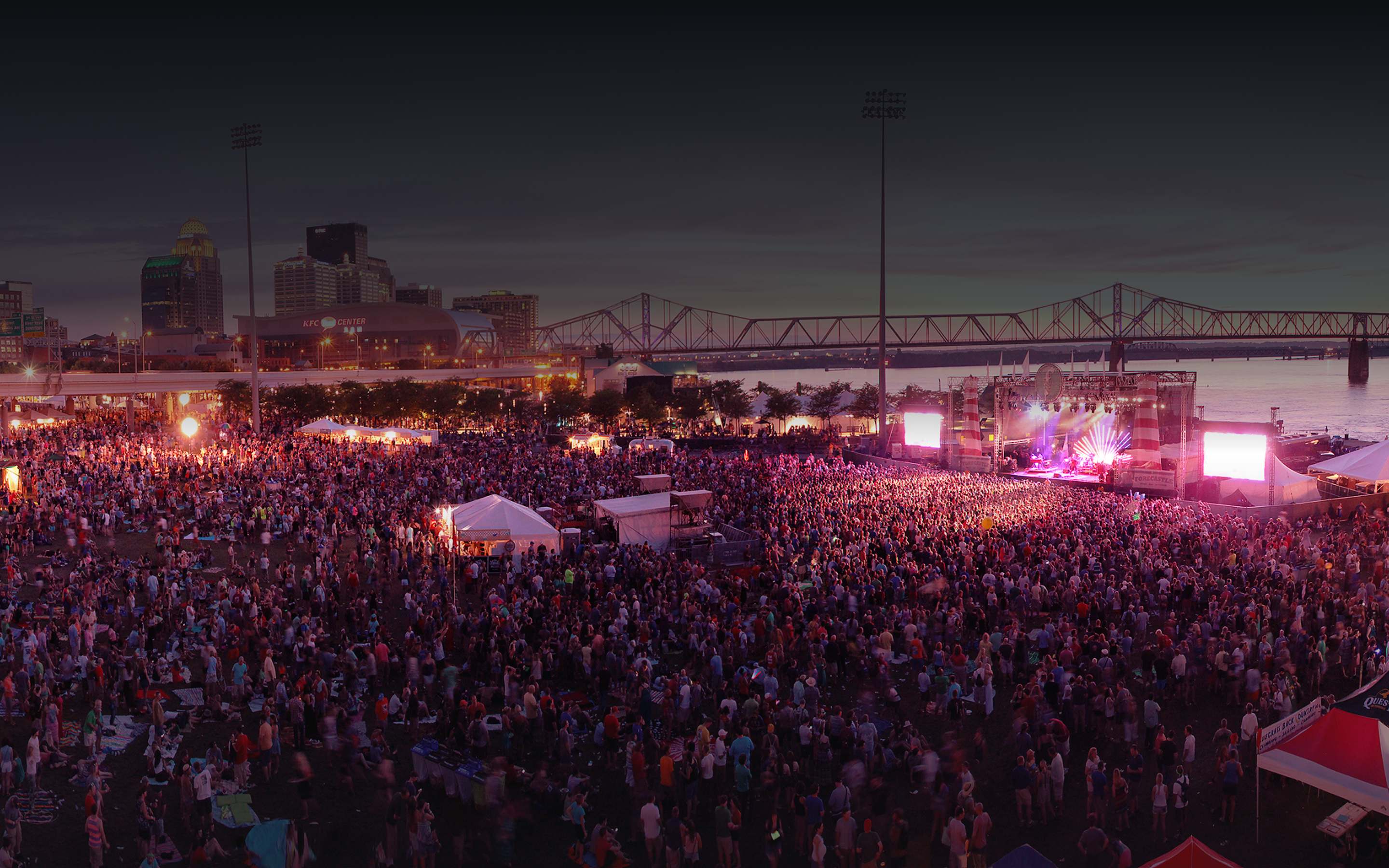 Large nighttime festival crowd on the Louisville riverfront with the downtown skyline, KFC Yum Center, and bridge behind under pink stage lighting