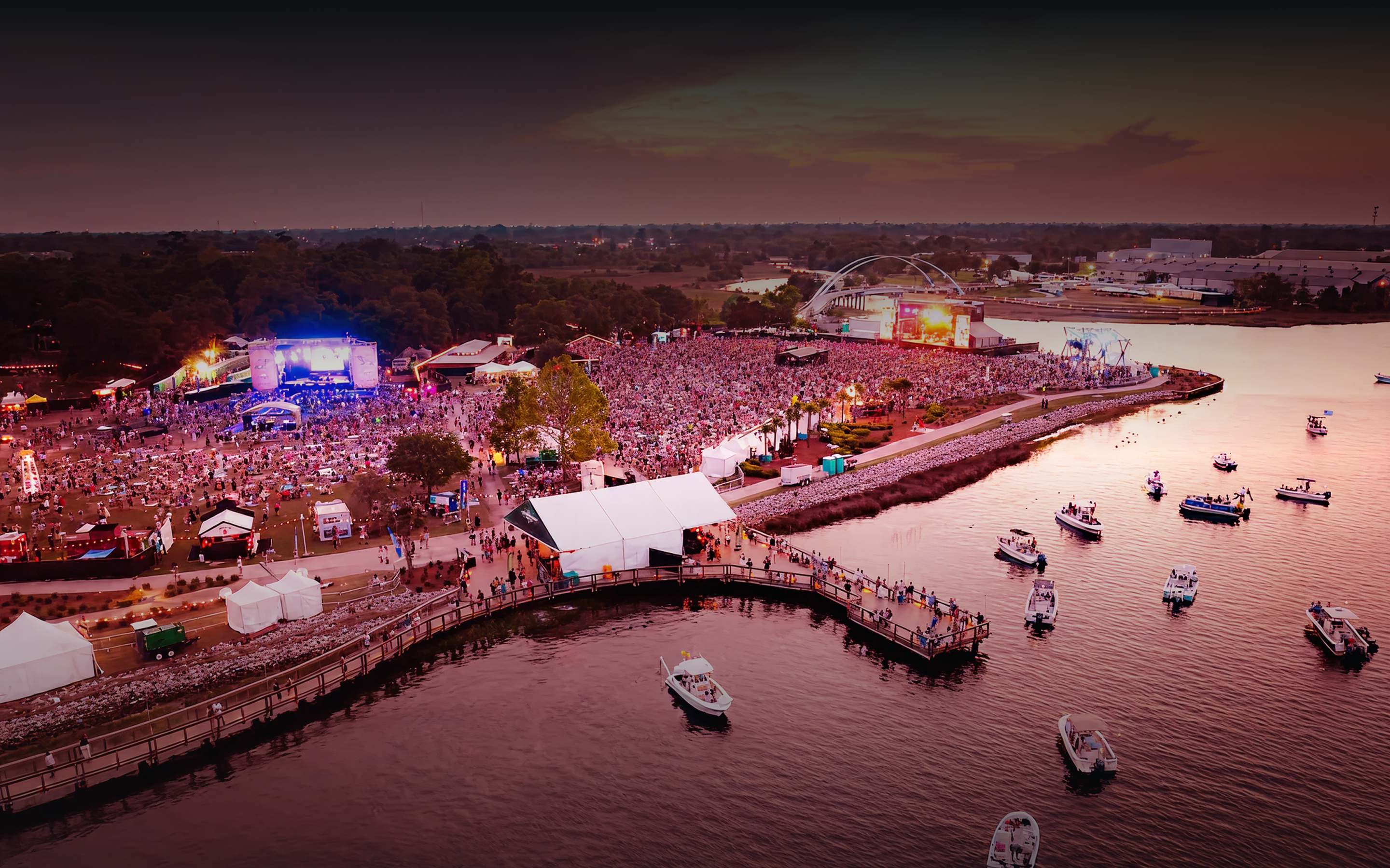 Aerial view of a waterfront music festival at sunset with two lit stages and a crowd along the pier