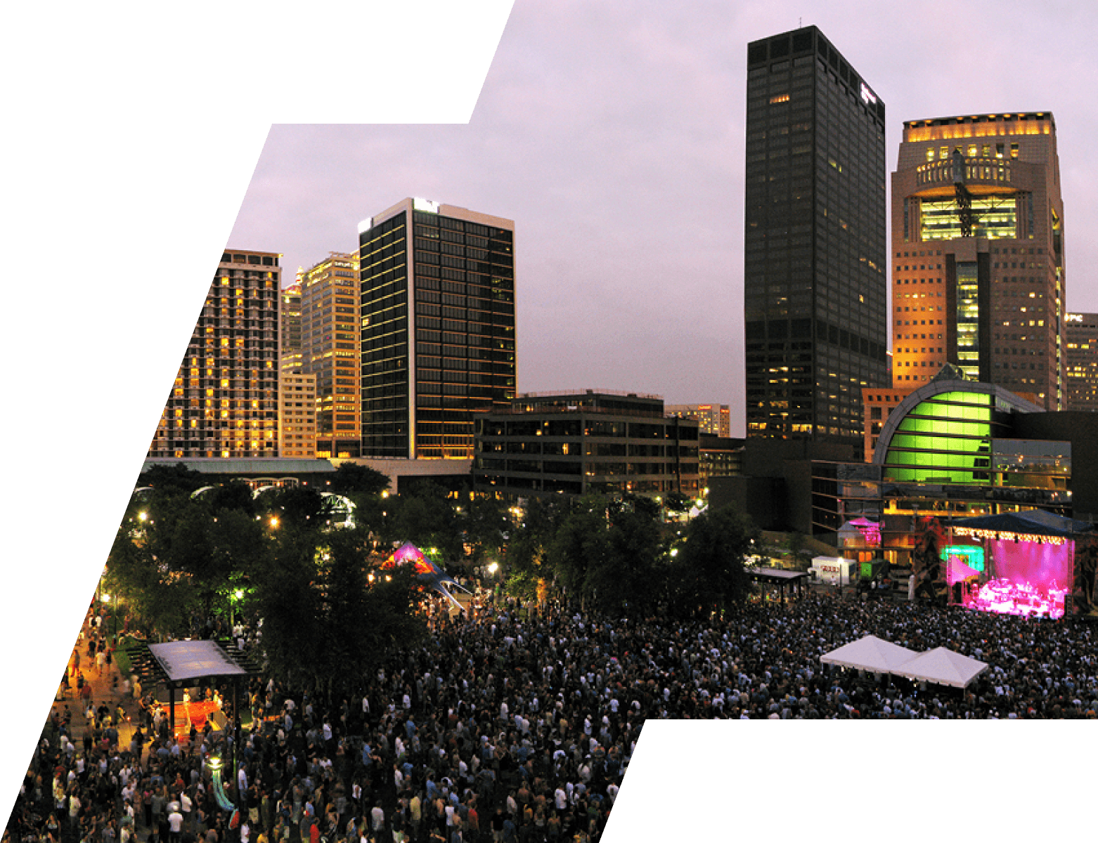 Downtown city skyline at dusk above a packed festival crowd in a riverfront park