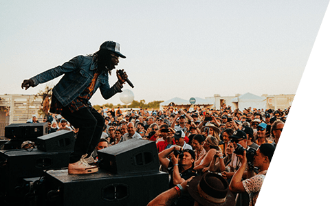 Festival performer on stage monitors with a mic facing the crowd at sunset