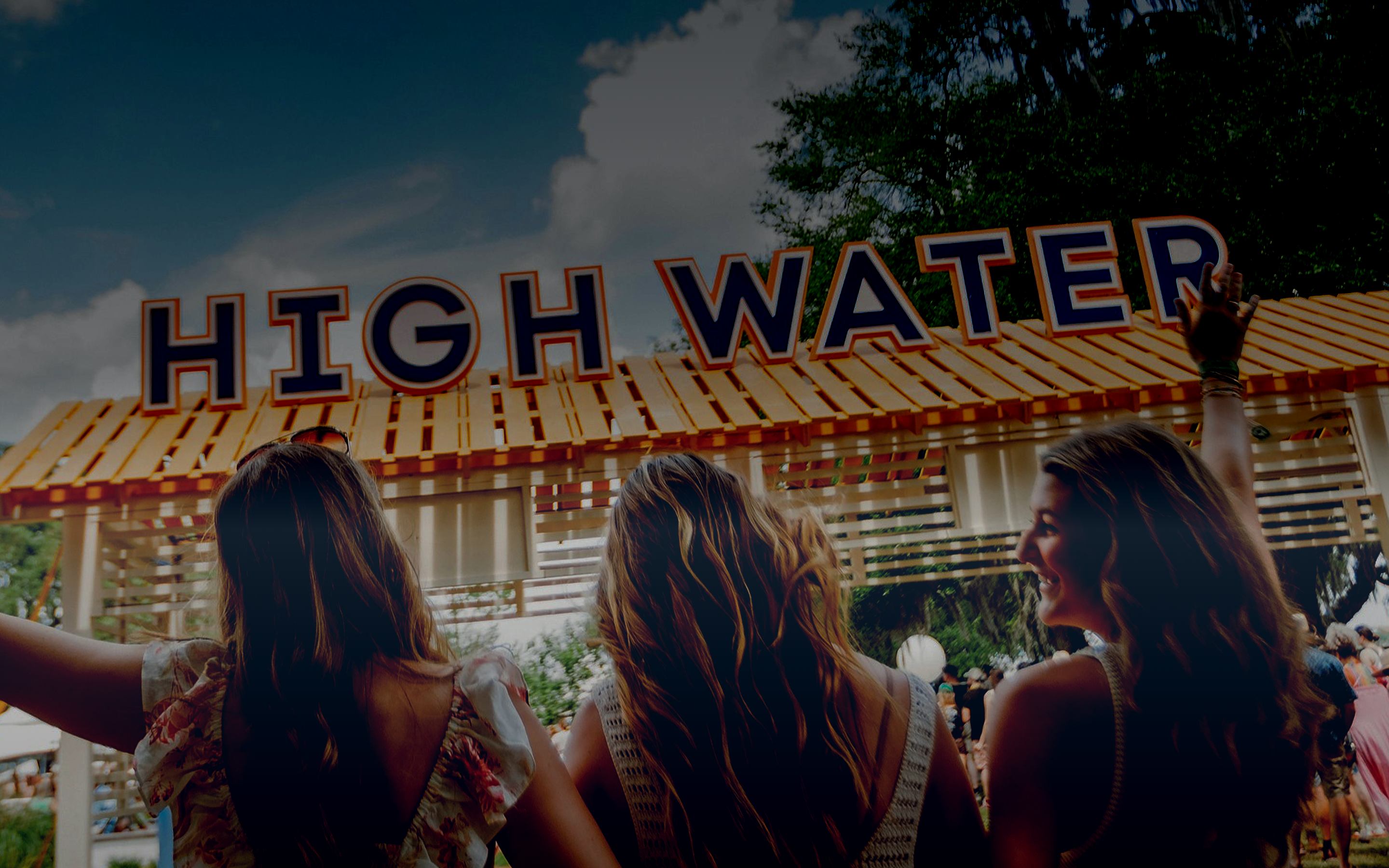 Three friends looking up at a large wooden festival sign under a blue sky