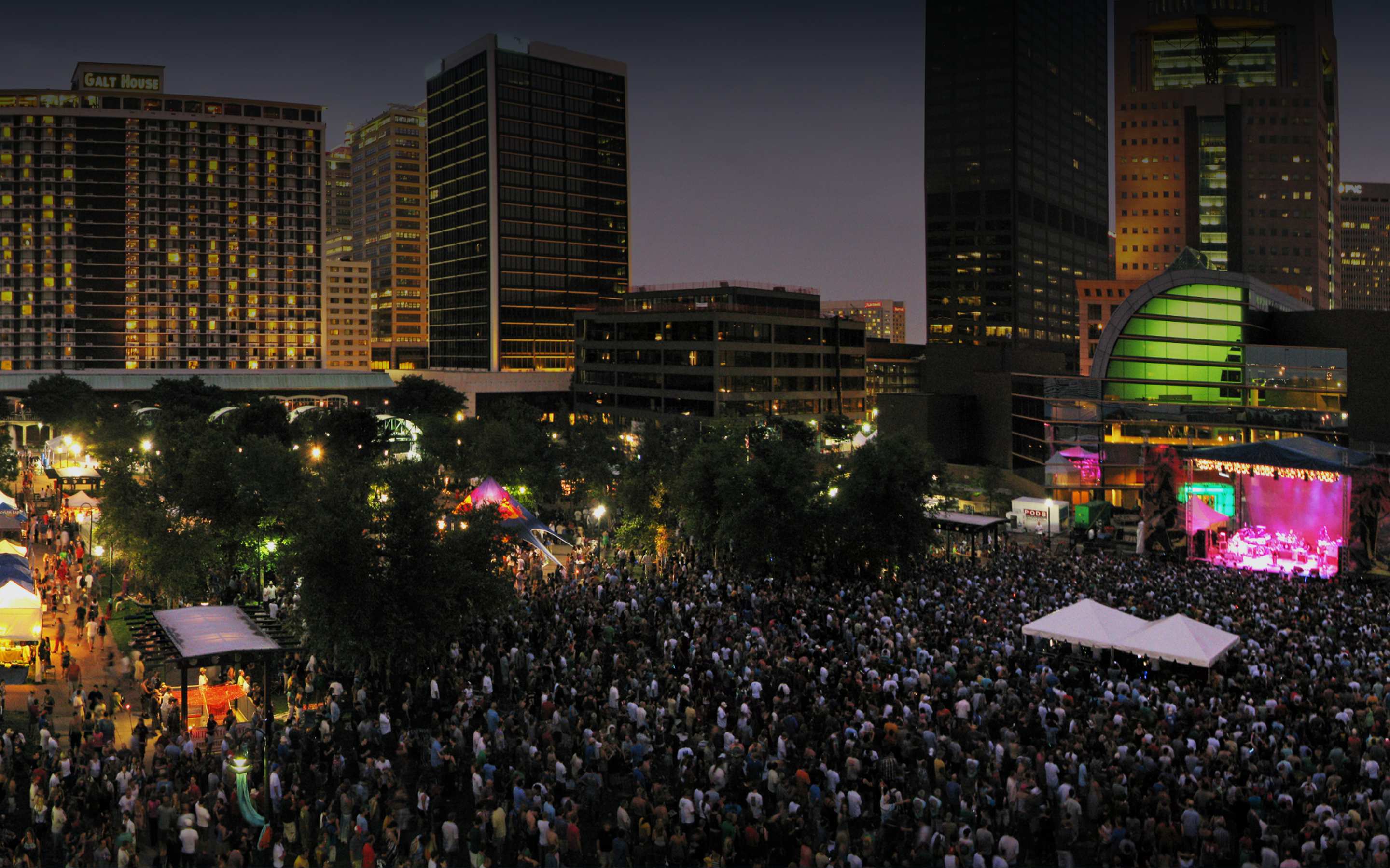 Large nighttime festival crowd in a downtown park facing a pink-lit main stage with the city skyline lit behind