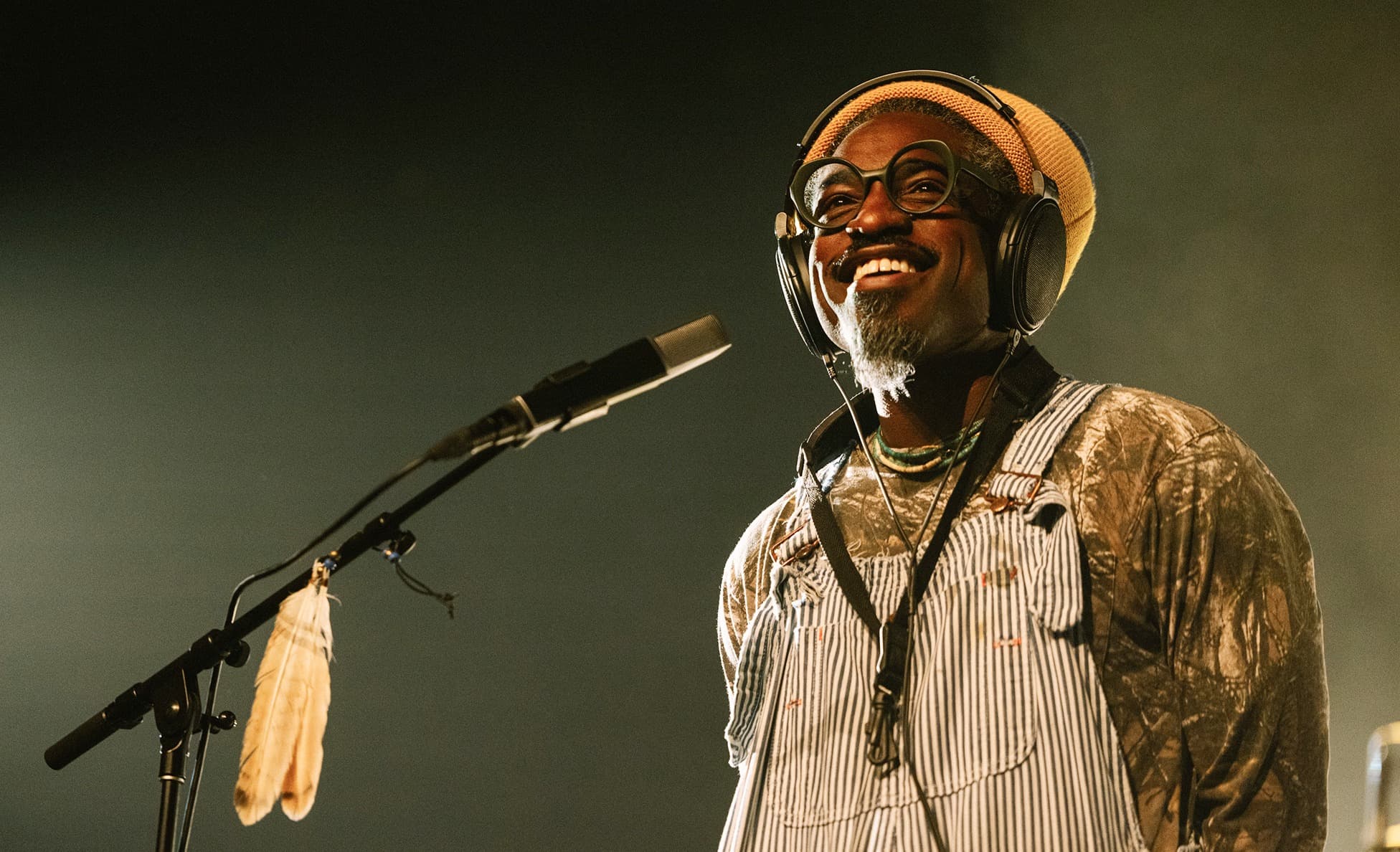 Smiling performer on stage wearing headphones, round glasses, a beanie, and striped overalls