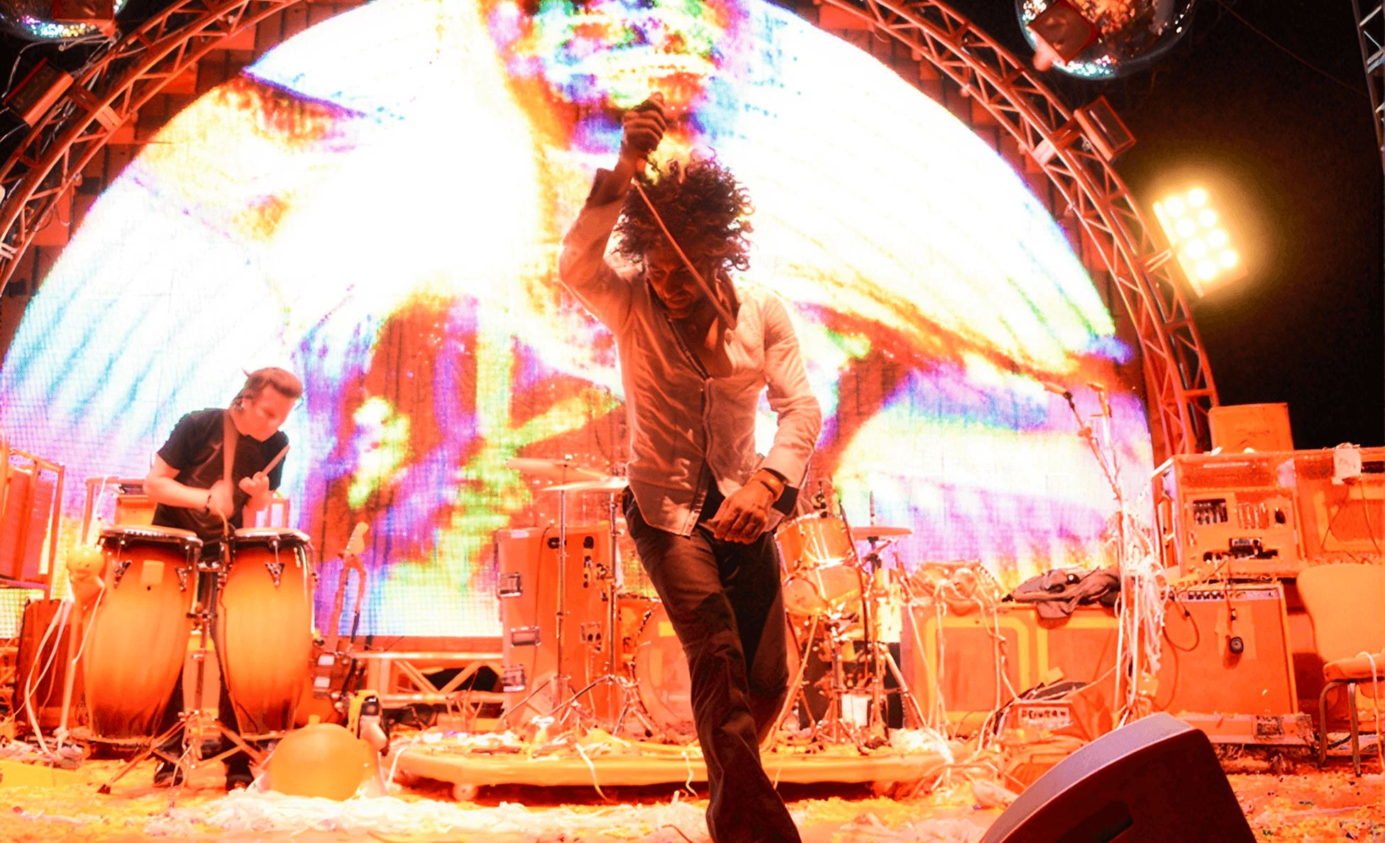 Frontman dancing on stage beside a percussionist in front of a psychedelic domed LED screen with confetti