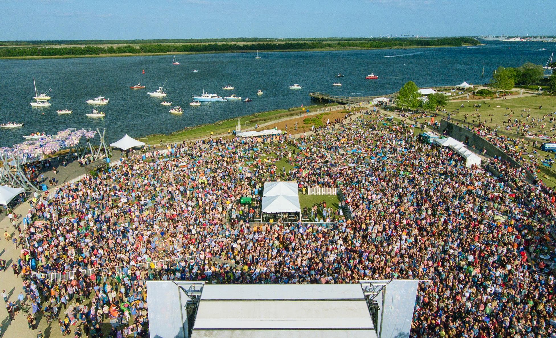 Aerial view of a massive festival crowd on a waterfront lawn beside a harbor full of boats