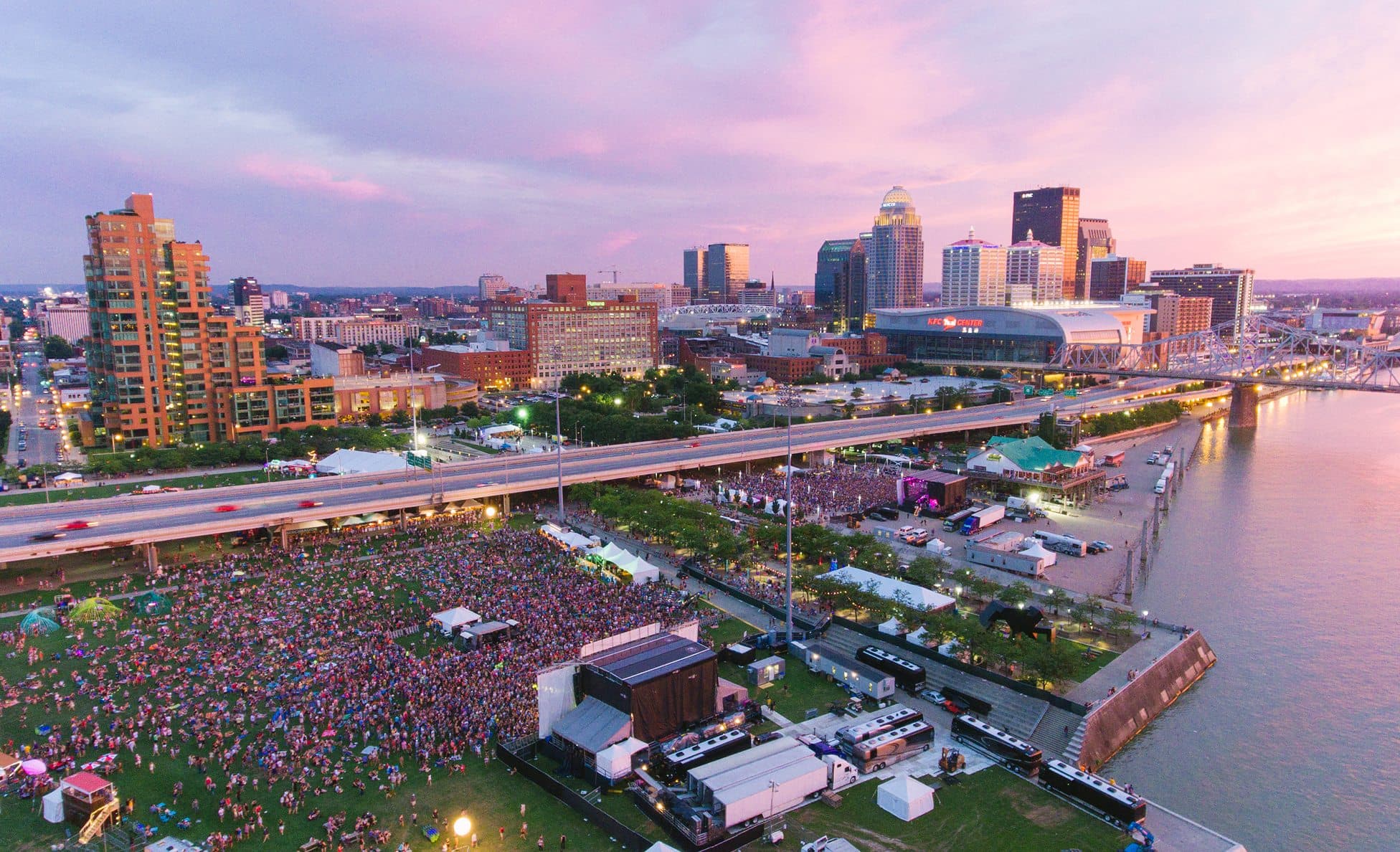 Aerial dusk view of a riverfront festival with the Louisville skyline behind the crowd