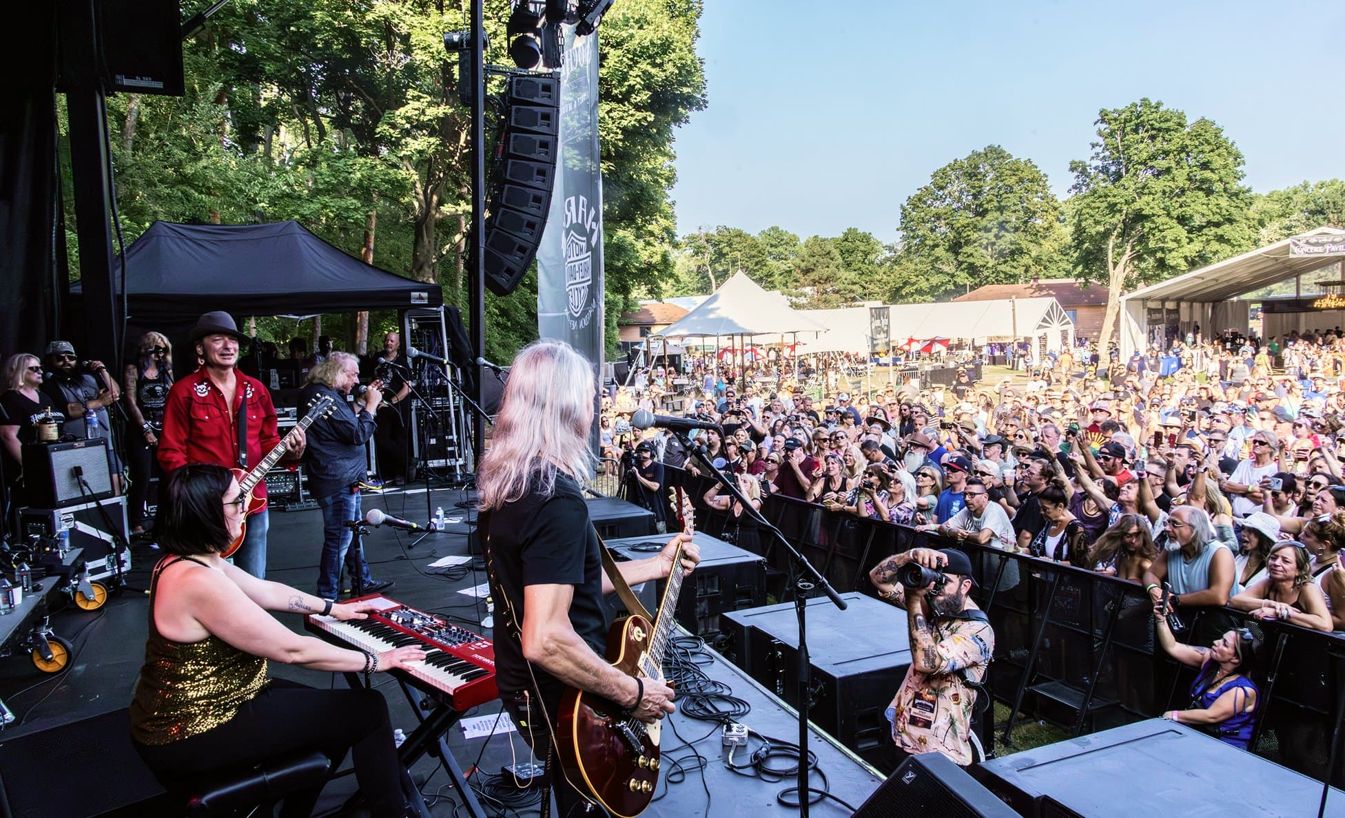 View from behind a band on stage looking out to a sunny crowd in a wooded amphitheater