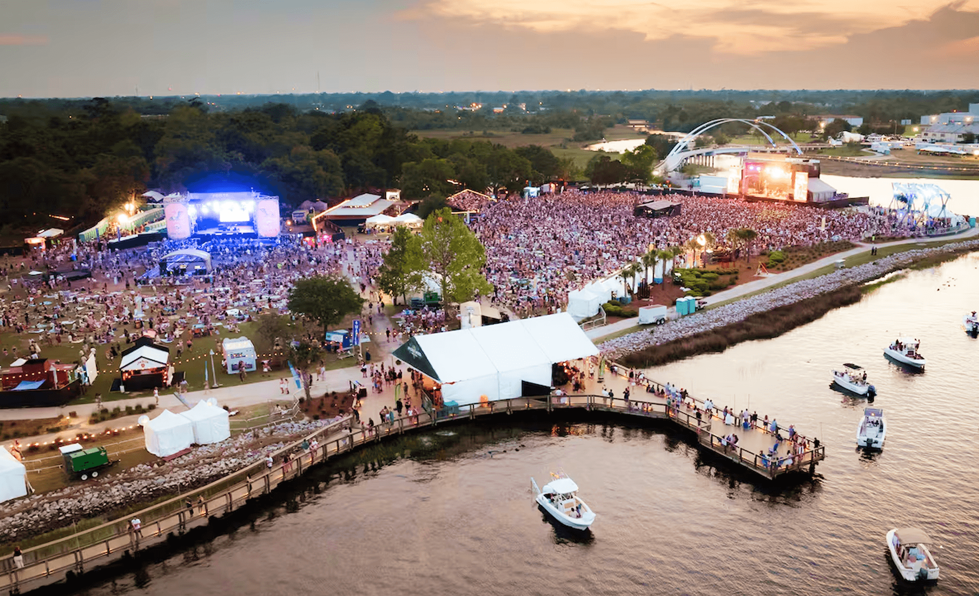Aerial sunset view of a waterfront festival with dual stages and a curving pier lined with boats