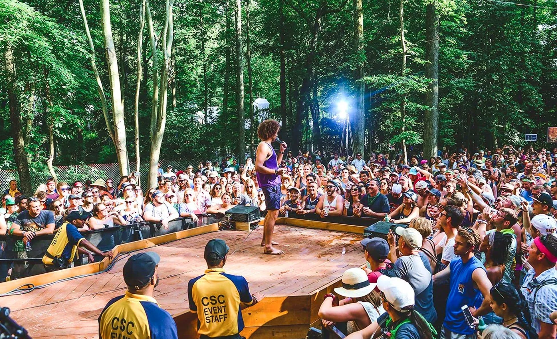 Performer in a purple tank top engaging a tight crowd on a wooden stage in the woods