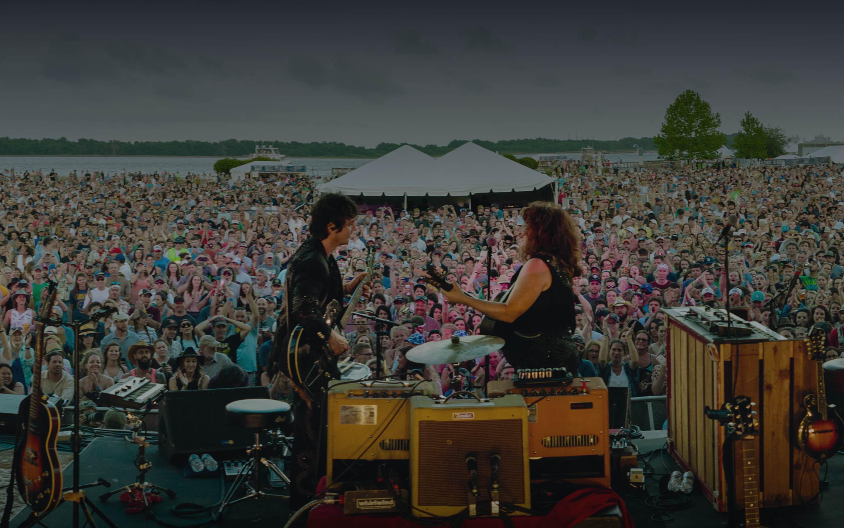 On-stage view of two musicians performing to a massive waterfront festival crowd under an overcast sky