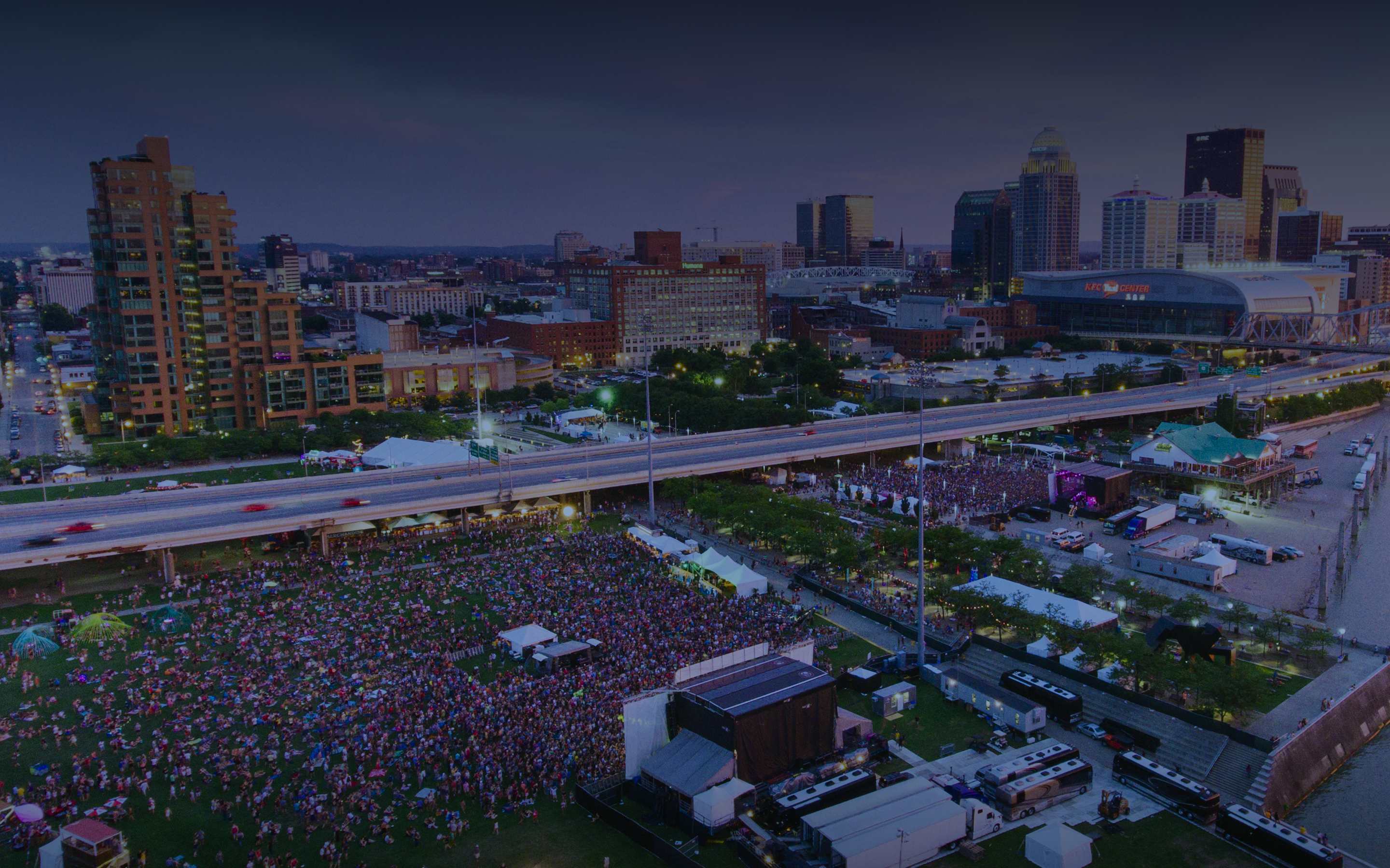 Aerial view of a waterfront festival at dusk with a large crowd, stages, and the downtown skyline behind