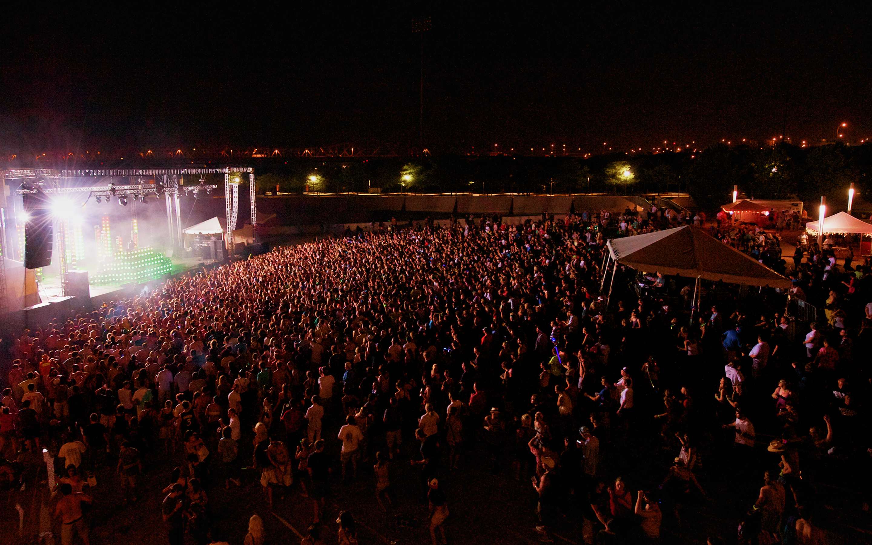 Aerial night view of a massive festival crowd facing a brightly lit main stage, with a lit riverfront bridge and city skyline in the background