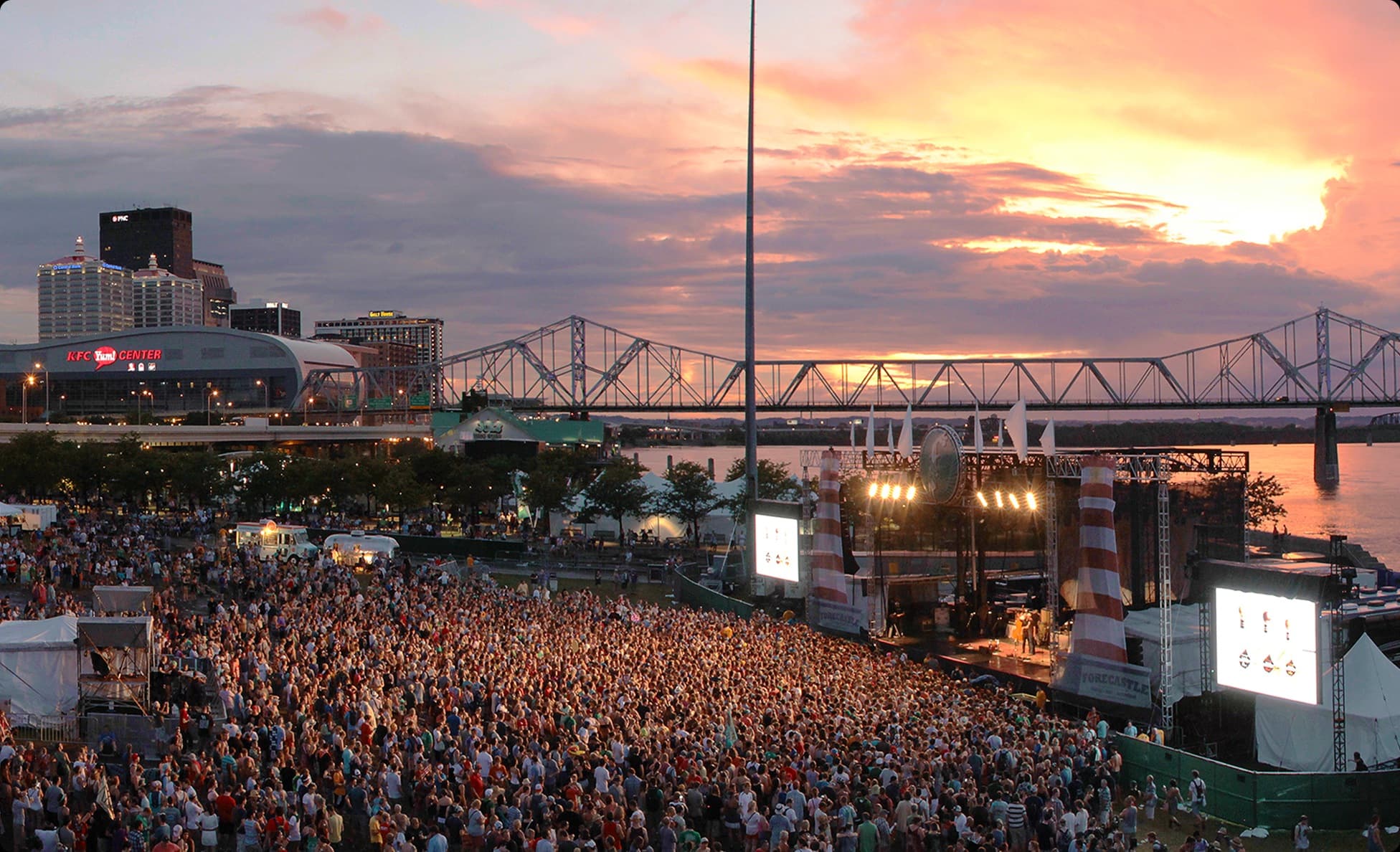 Waterfront festival main stage beneath a riverside sunset skyline