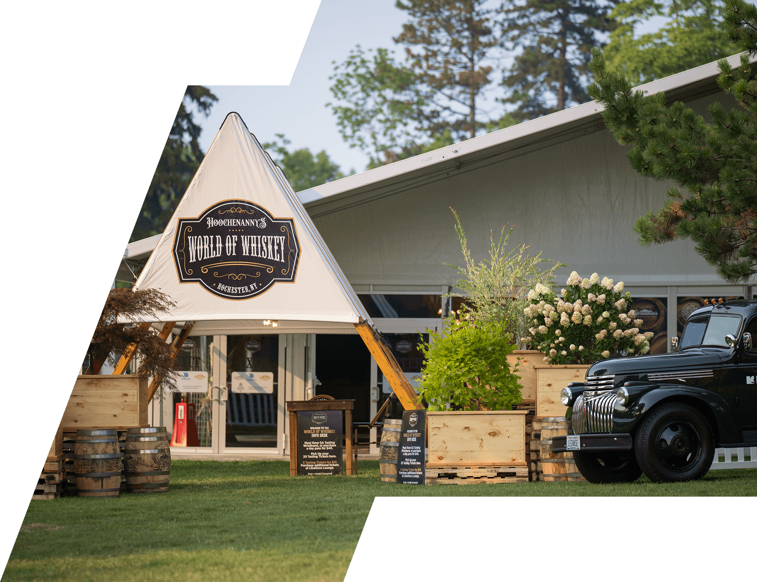 Hoochenanny's World of Whiskey A-frame tent activation with whiskey barrels, planters, and a vintage green pickup truck on a festival lawn
