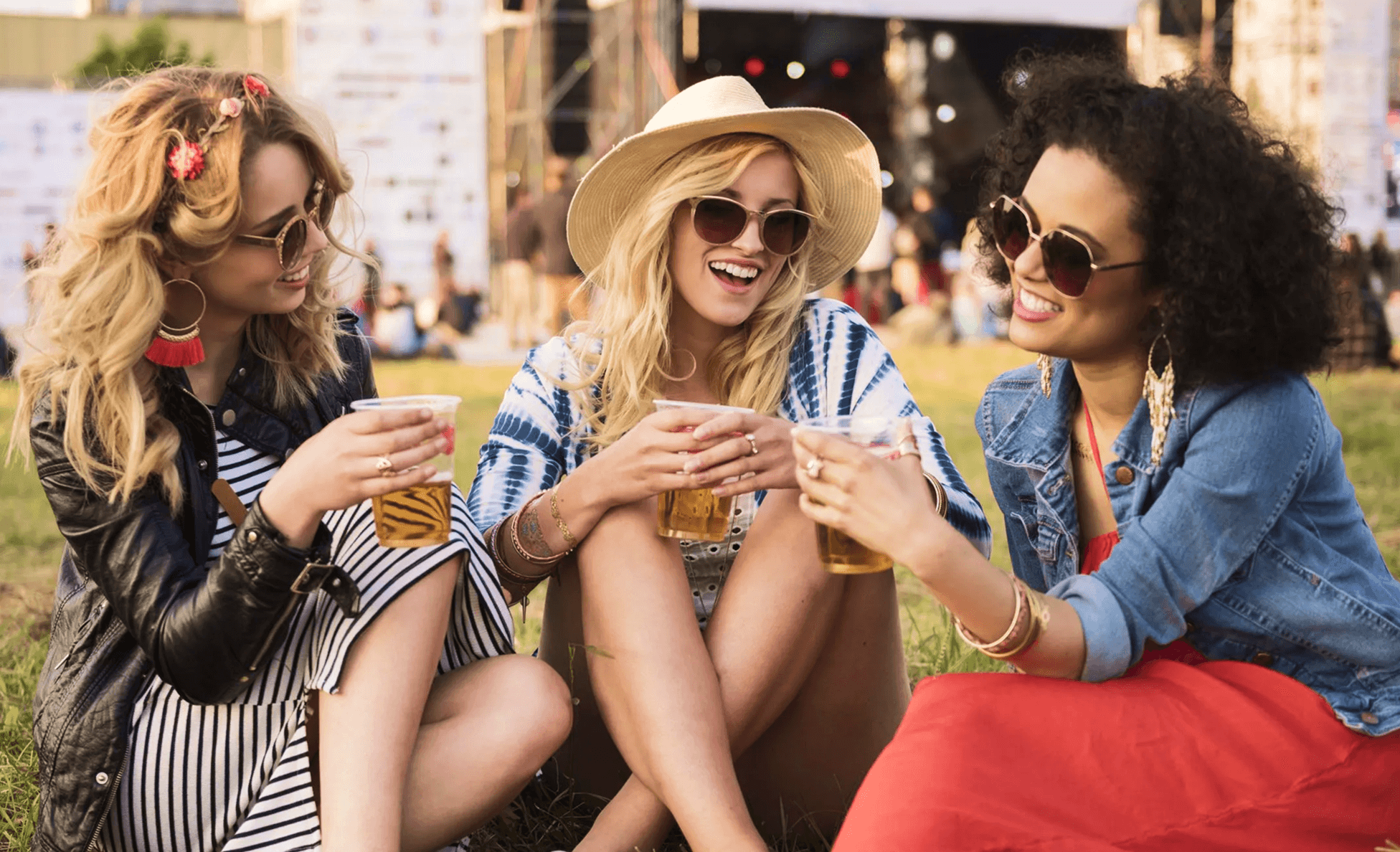 Three festival-goers in sunglasses laughing and toasting cups of beer on the lawn