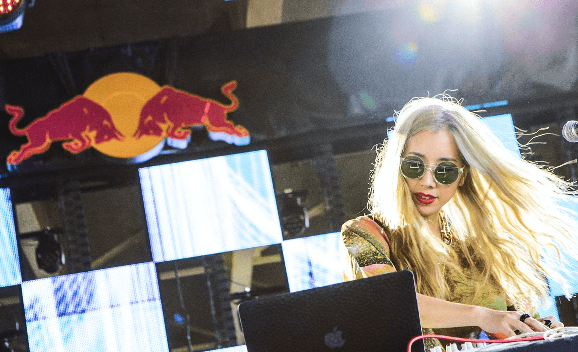 Blonde DJ in green round sunglasses performing under a Red Bull logo and video wall