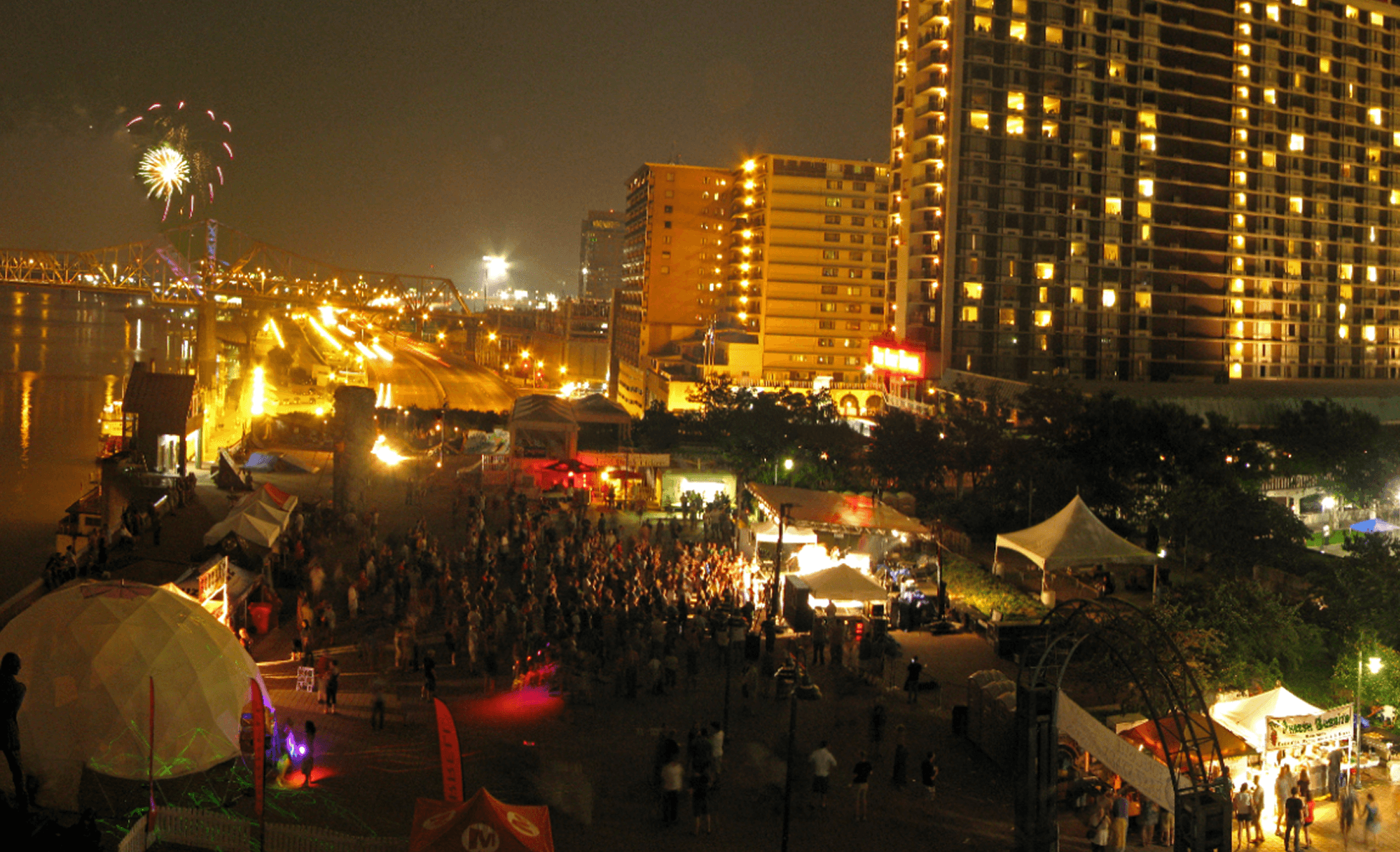Night aerial of a riverfront festival with fireworks bursting over a bridge and lit skyline