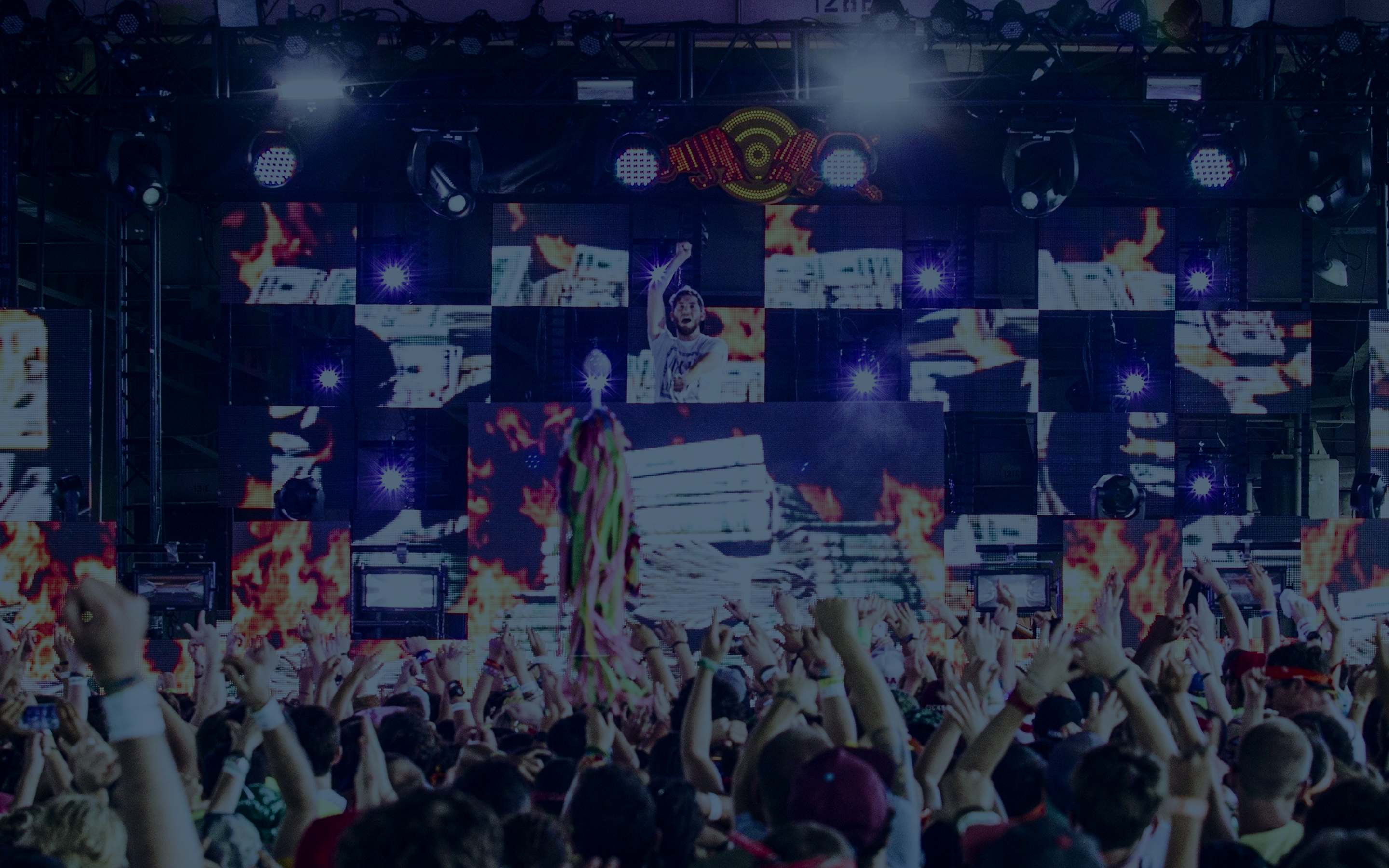 Festival crowd with hands raised in front of a massive LED main stage lit with flames and stage lighting at night