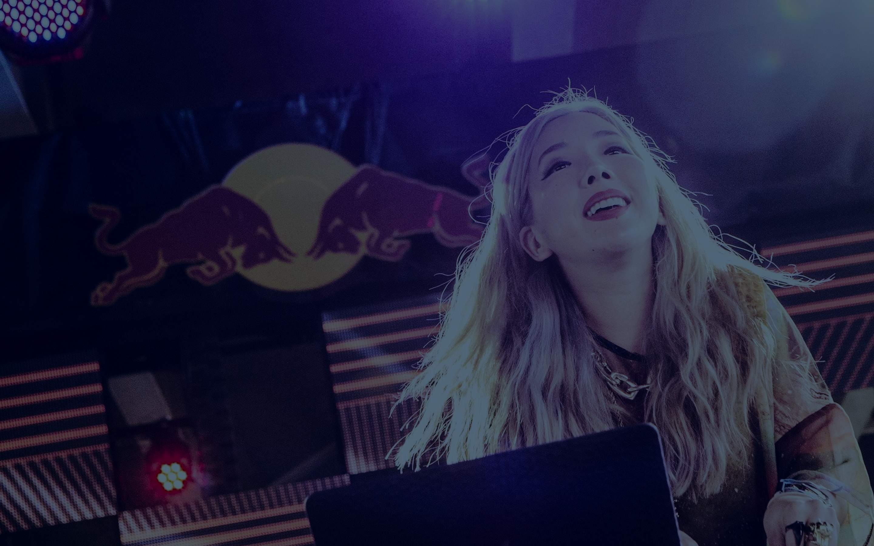 Blonde DJ looking up joyfully while performing behind a laptop at a Red Bull-sponsored festival stage